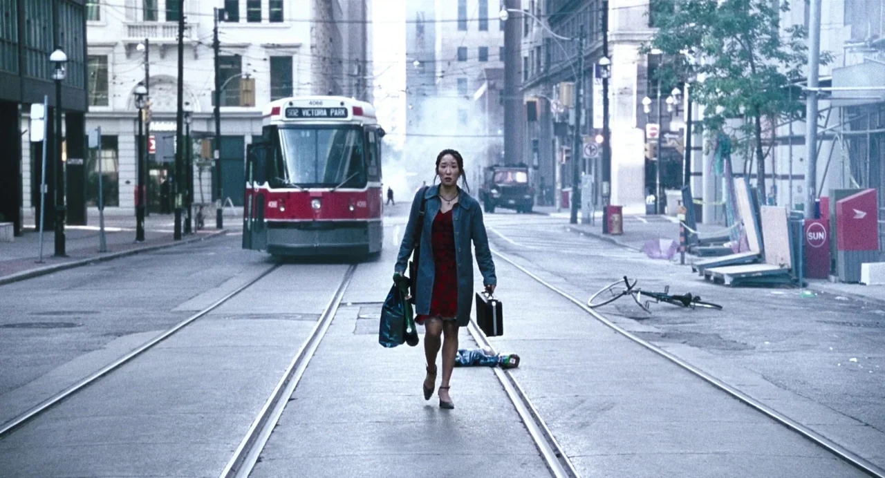 Last Night (1998) movie still: fullbody — An empty urban street with tram tracks features a large red streetcar behind the woman. A woman walks…