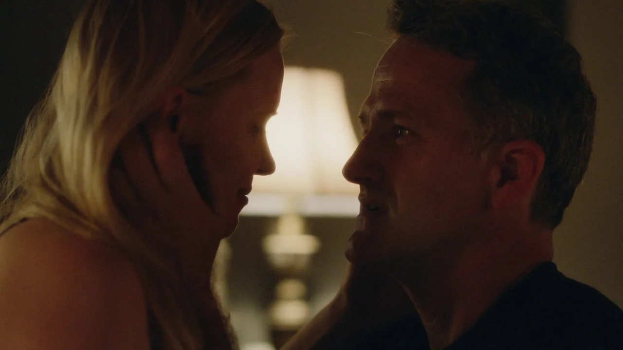 Saint Frances (2019) movie still: closeup — A dark, dimly lit room contains a glowing table lamp between two people. A man and a woman stand face to…