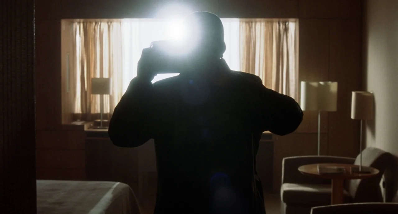 Femme Fatale (2002) movie still: medium — A hotel room with wood-paneled walls and a chair near a large bright window. A person standing in the…