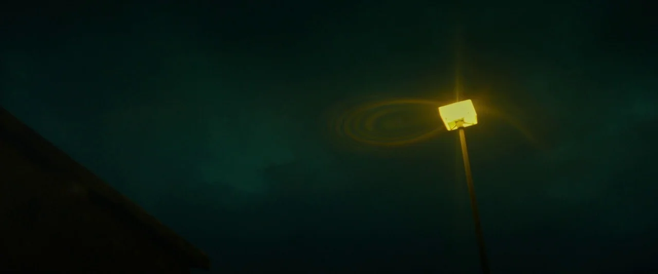 Kissing Candice (2017) movie still: establishing — A dark night sky frames a single glowing streetlamp with a yellow fixture. A faint spiral-shaped…