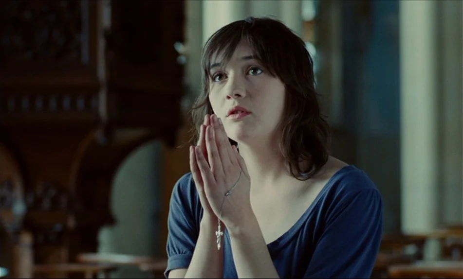 Hadewijch (2009) movie still: medium — A stone cathedral interior features blurred wooden pews and architectural arches in the background. A young…