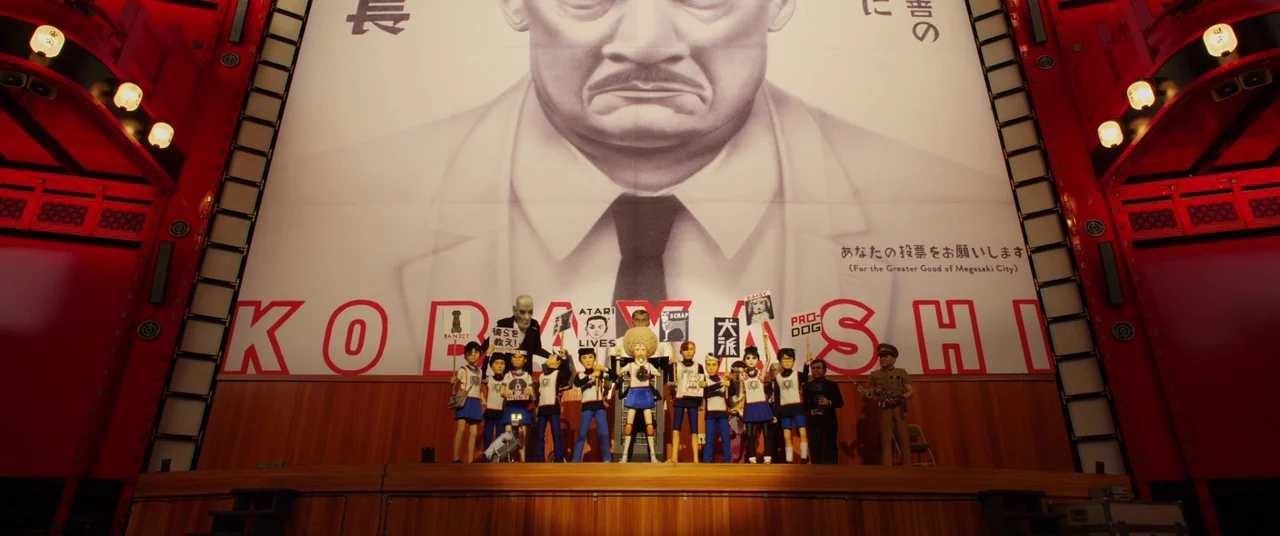 Isle of Dogs (2018) movie still: wide — A group of puppets stands on a wooden stage before a large campaign banner. The banner features a giant…