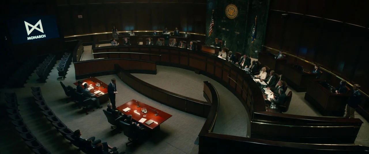 Godzilla: King of the Monsters (2019) movie still: wide — A spacious legislative chamber features curved wooden desks and a large video screen…