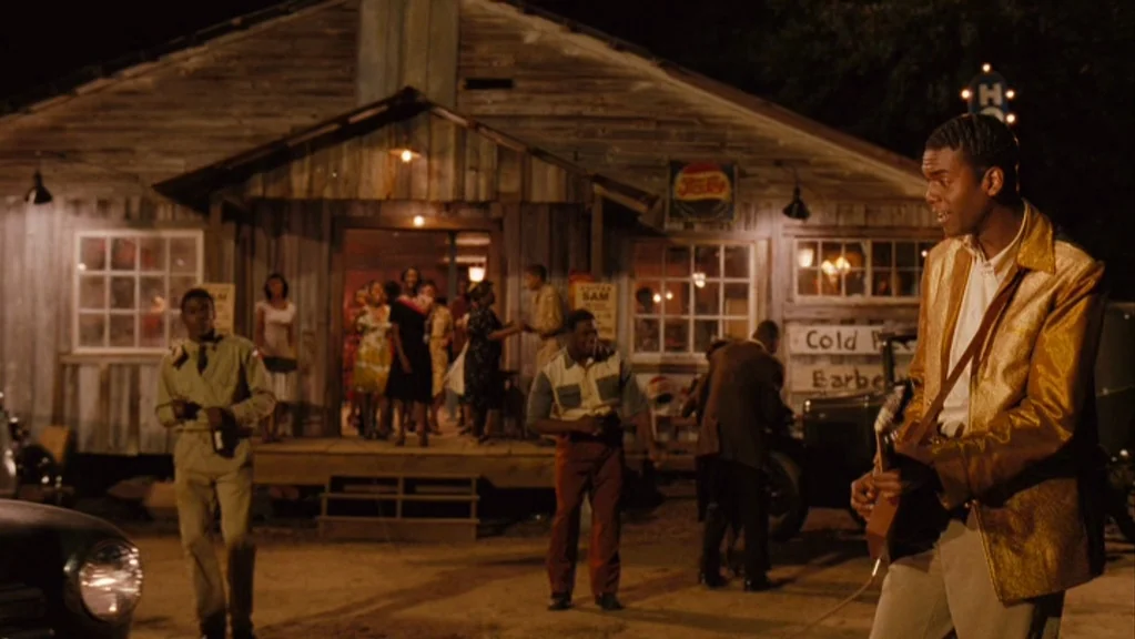 Honeydripper (2007) movie still: wide — A wooden building with a front porch serves as a gathering place at night. Several people interact on the…