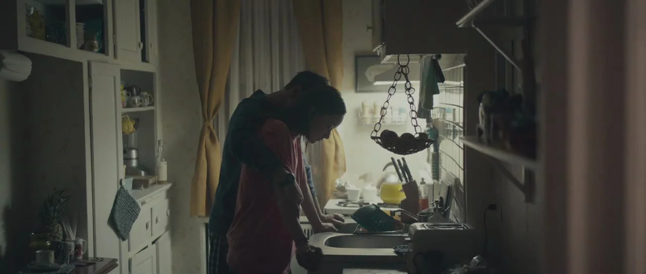Meadowland (2015) movie still: medium — A cluttered kitchen with white cabinets, yellow curtains, and a sink area. A man embraces a woman from…