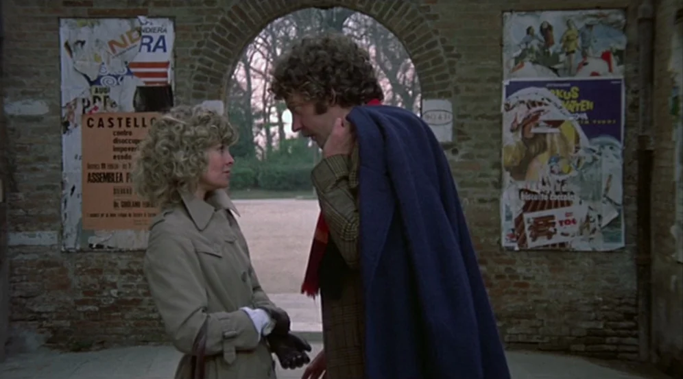 Don’t Look Now (1973) movie still: medium — A rustic brick archway is flanked by two weathered posters on aged walls. A woman and a man in coats…