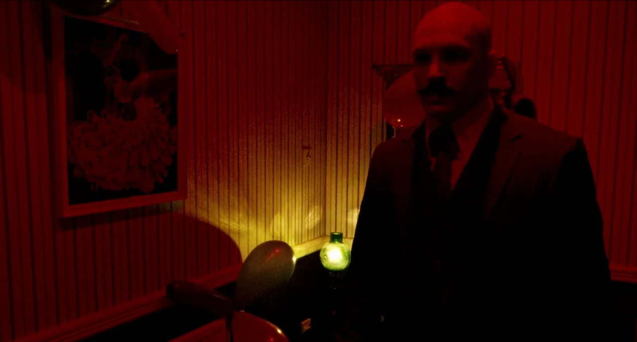 Bronson (2008) movie still: medium — A dimly lit hallway with vertical striped wallpaper features party balloons on the floor. A man wearing a suit…