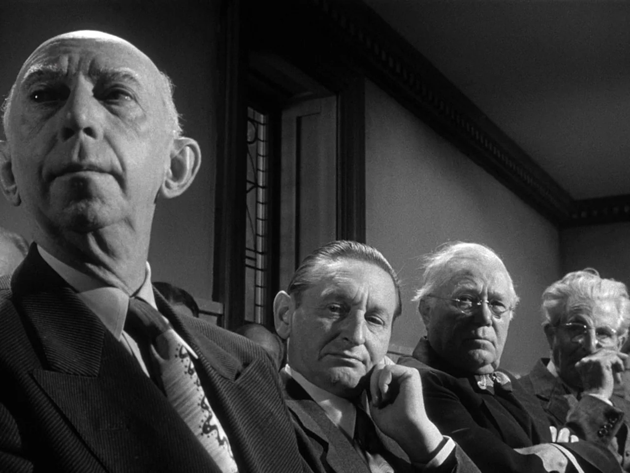 The Prowler (1951) movie still: medium — A dark wooden room with paneled walls and a tall, narrow window. Four men in suits sit in a row and look…