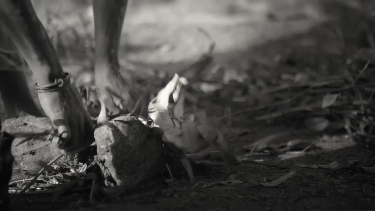 Faya Dayi (2021) movie still: closeup — An outdoor ground surface is covered in dirt, scattered dry leaves, and small stones. Hands manipulate…