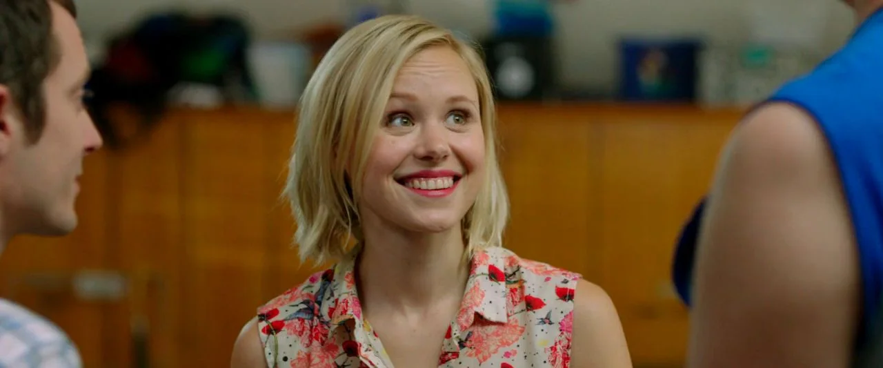Cooties (2014) movie still: medium — An interior space with orange wooden lockers lining the background wall. A smiling woman stands between two…