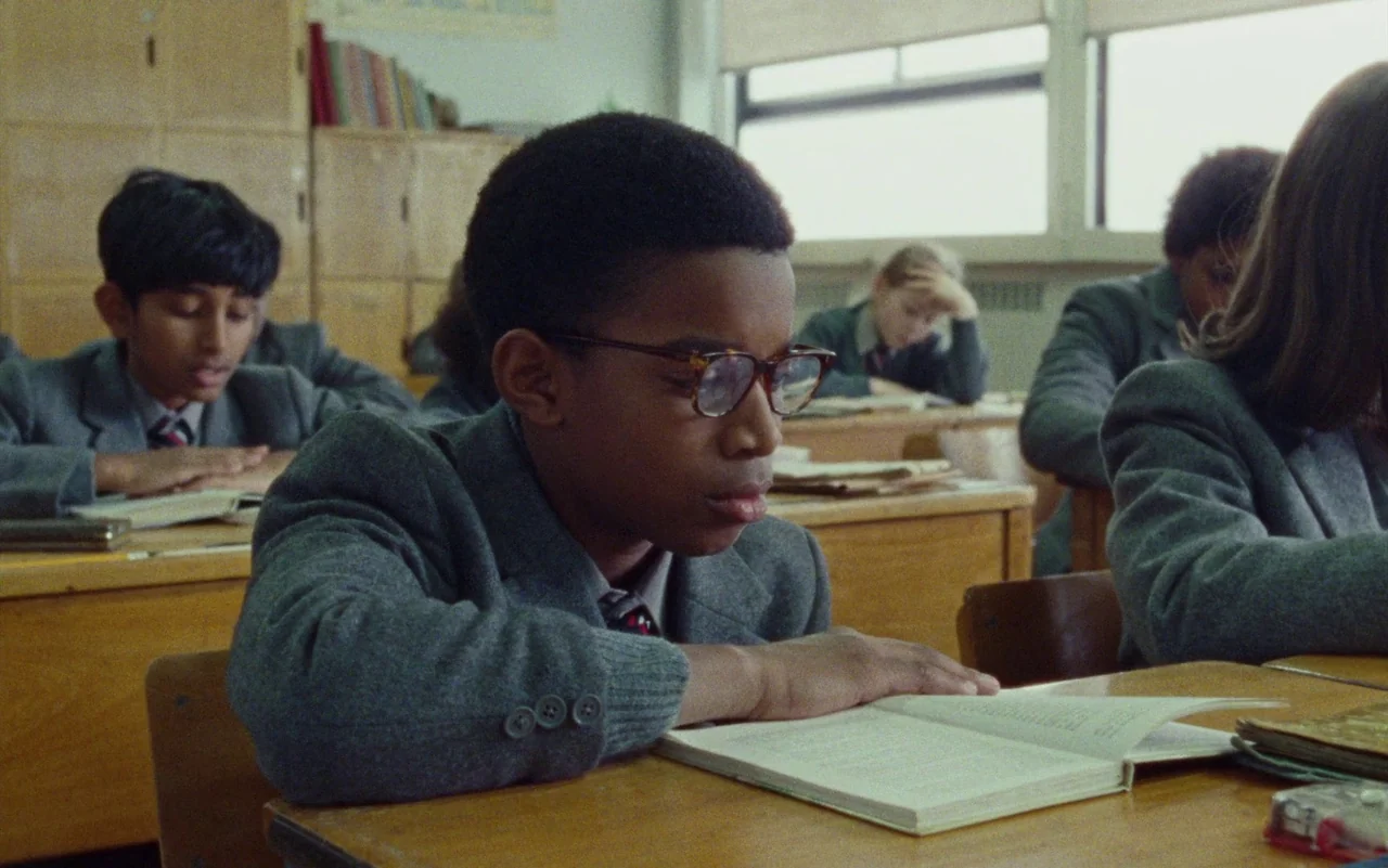 Education (2020) movie still: medium — A school classroom with rows of wooden desks and students in grey school blazers. A young boy with glasses…