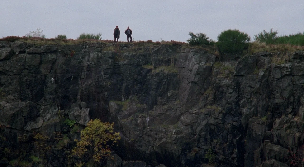 Shallow Grave (1994) movie still: wide — A vertical rock face rises sharply from the ground to a grassy ridge top. Two small figures stand…