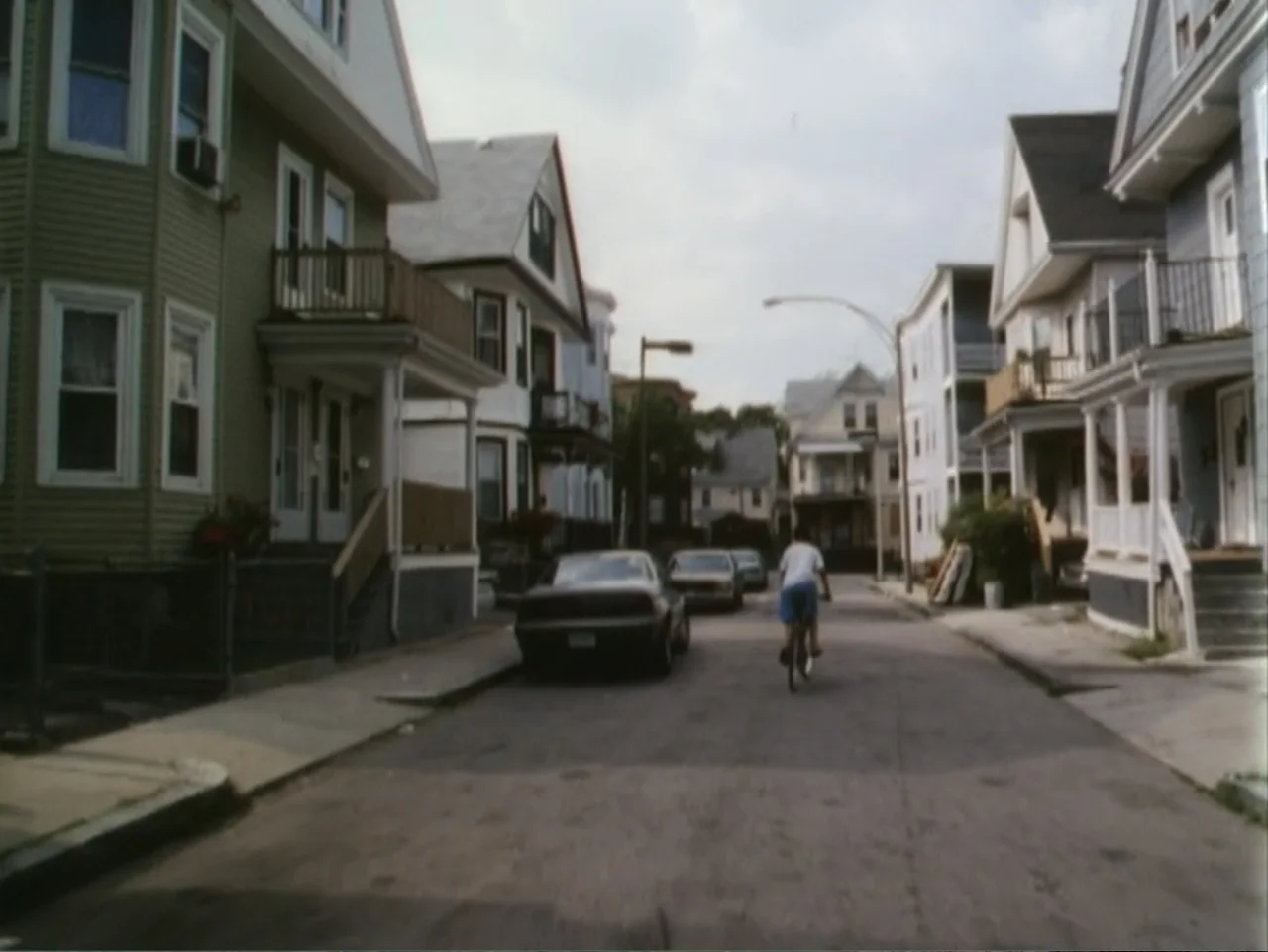 5 American Handguns - 5 American Kids (1995) movie still: wide — A residential street lined with multi-story wood-frame houses and parked cars. A…