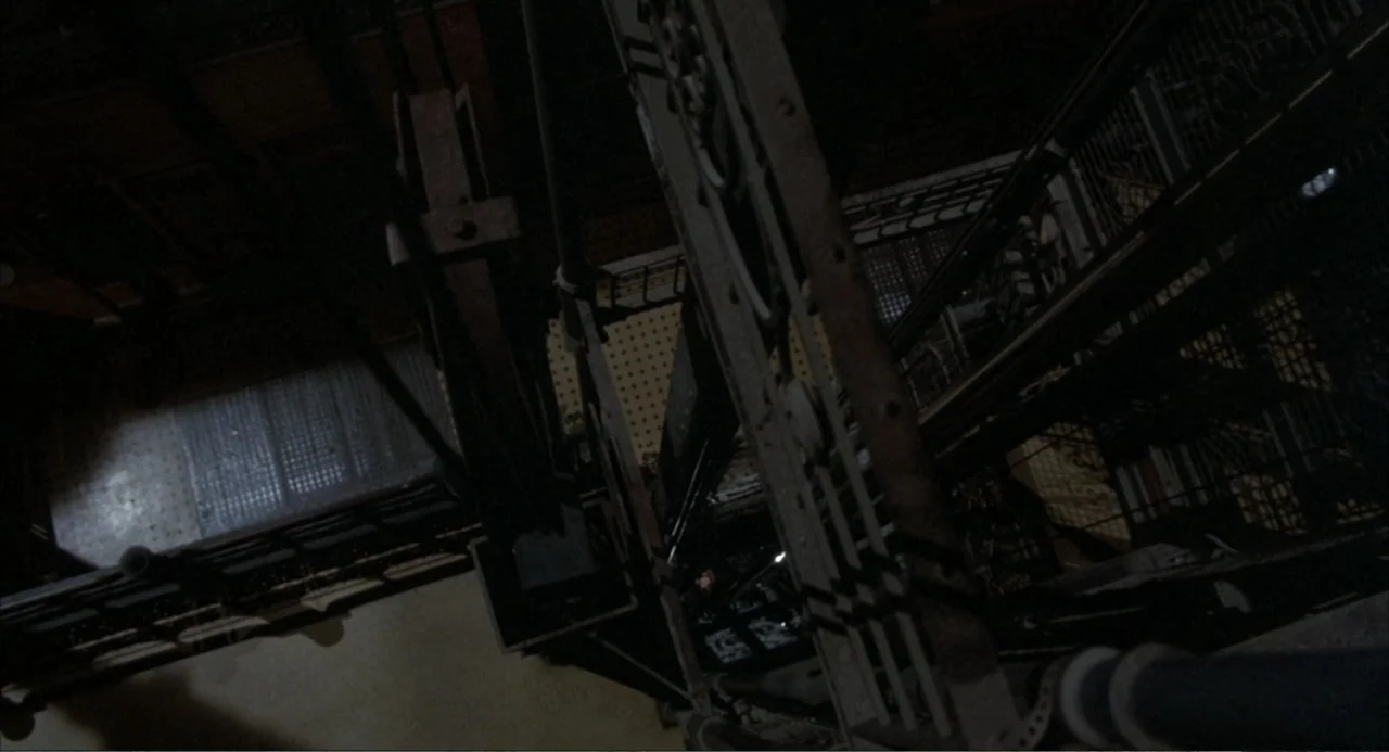 Murphy's Law (2009) movie still: establishing — An industrial elevator shaft features thick steel cables and a lattice metal cage structure. The…