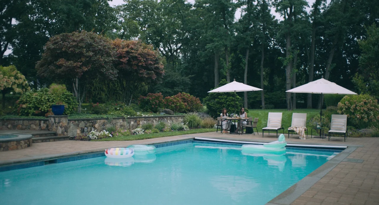 She Said (2022) movie still: wide — A stone patio area beside a swimming pool features lounge chairs and dining furniture. Two women sit at a table…