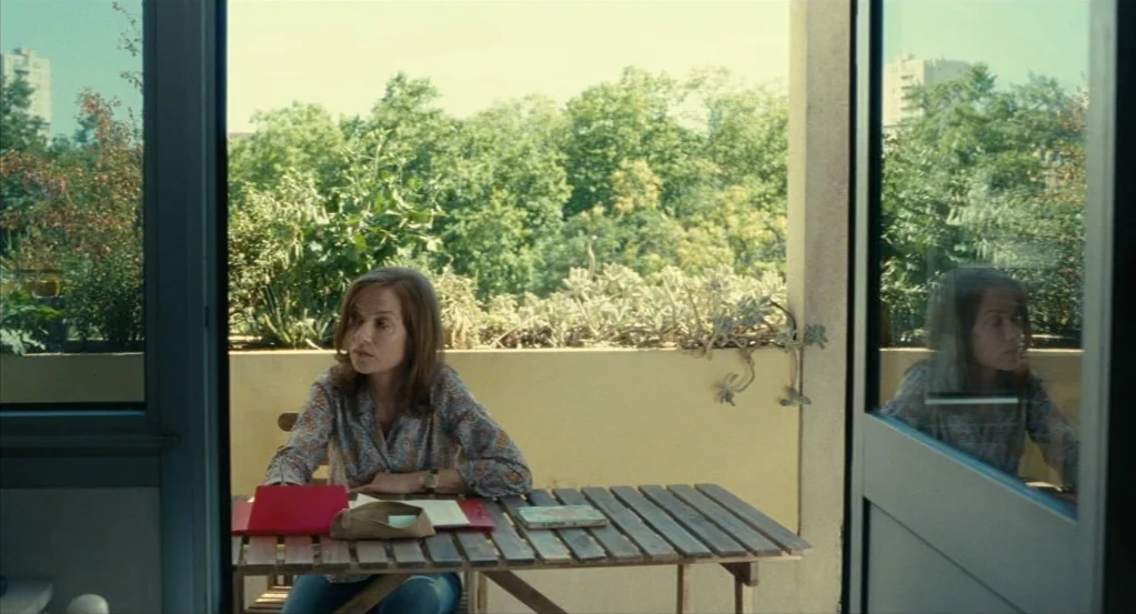 Things to Come (2016) movie still: medium — A sunny outdoor balcony with a slatted wooden table overlooks lush green trees. A woman sits at the…