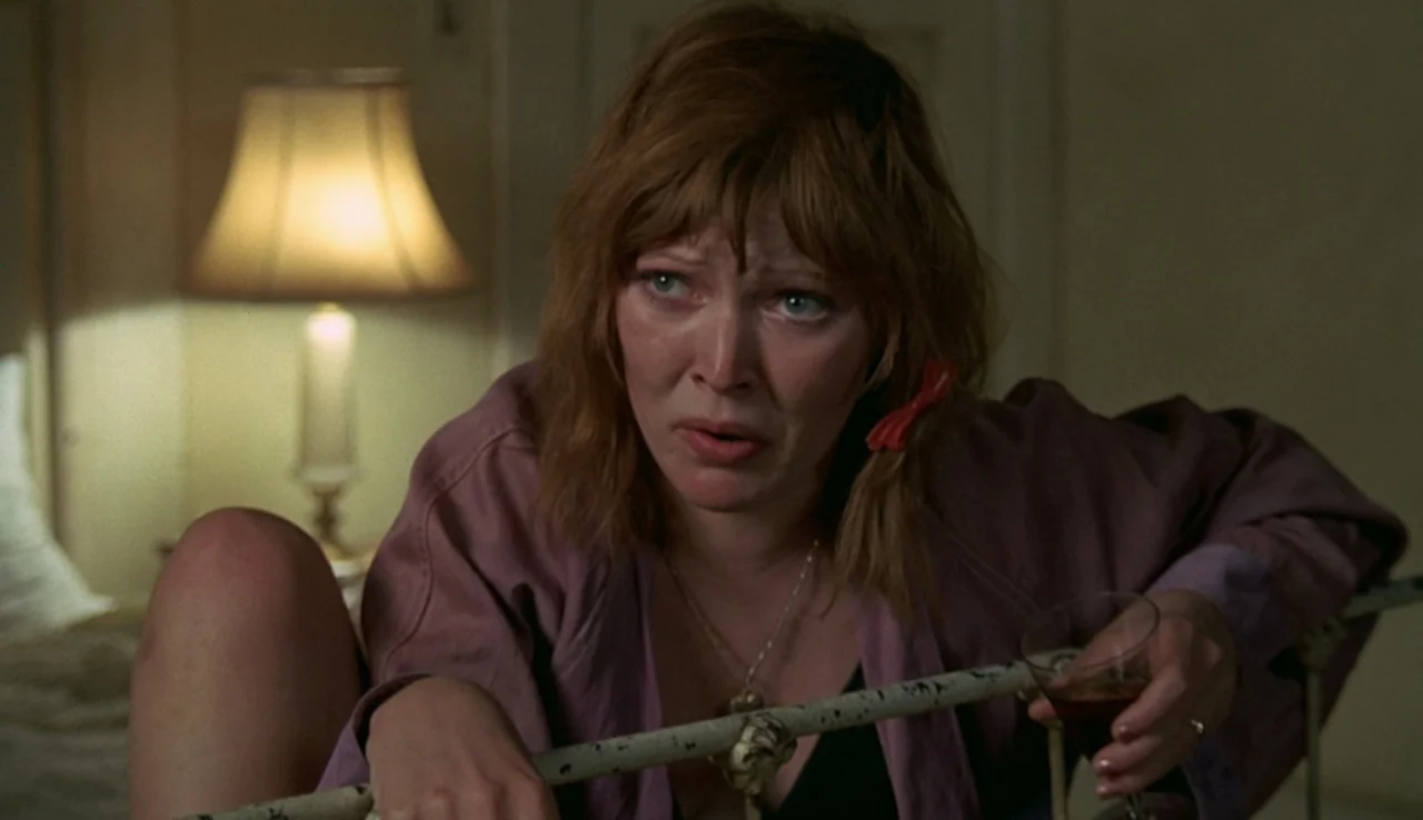 Fat City (1972) movie still: medium — A dimly lit room contains a white metal headboard and a bedside lamp. A woman sits on the bed holding a glass…