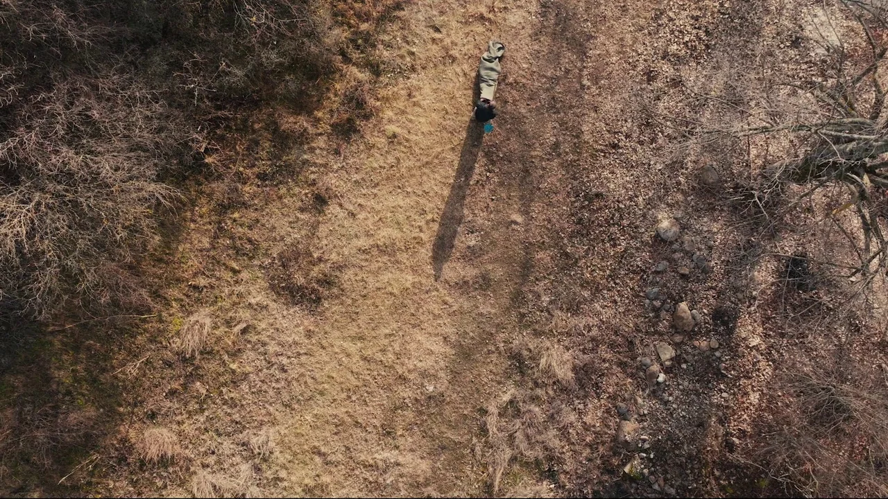 Helll Hole (2024) movie still: wide — A dry open field with patchy grass and scattered stones under bright sunlight. A person drags a large, folded…