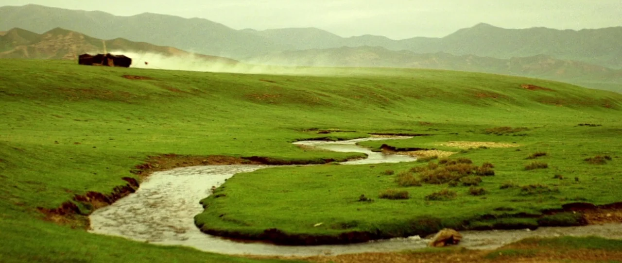 Horse Thief movie still: establishing — A vast green rolling prairie with a winding stream in the foreground. Low mountains are visible on the…