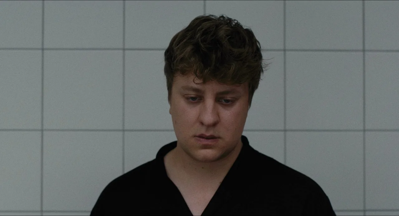 Afire (2023) movie still: closeup — A person stands in front of a white grid-patterned tiled wall. The man in a black shirt looks downward with a…
