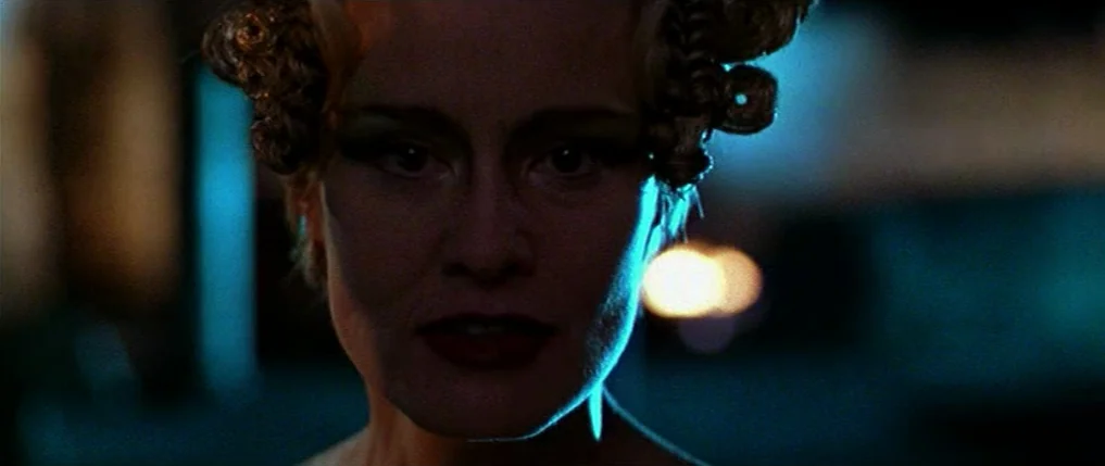 Titus (1999) movie still: closeup — A dark, dimly lit environment features a woman with elaborate blonde hair rolls. The subject stands illuminated…