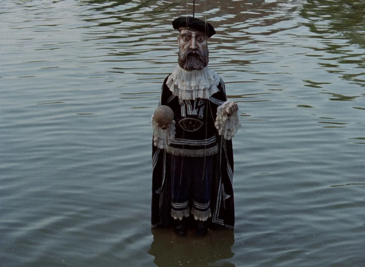 Faust (1994) movie still: fullbody — A marionette puppet stands vertically in calm, murky water. The puppet wears historical attire while holding a…