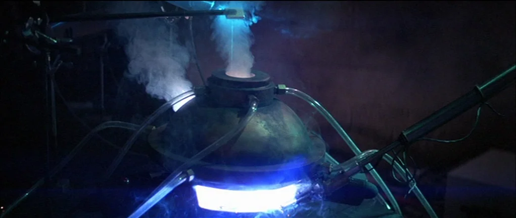 Demon Seed (1977) movie still: closeup — A dark laboratory features a complex dome-shaped metal machine with connected clear tubes. Thick steam…