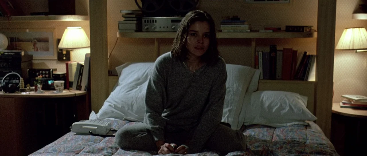 Opera (1987) movie still: medium — A furnished interior space with books on shelves and dual bedside lamps. A woman in a grey outfit sits…