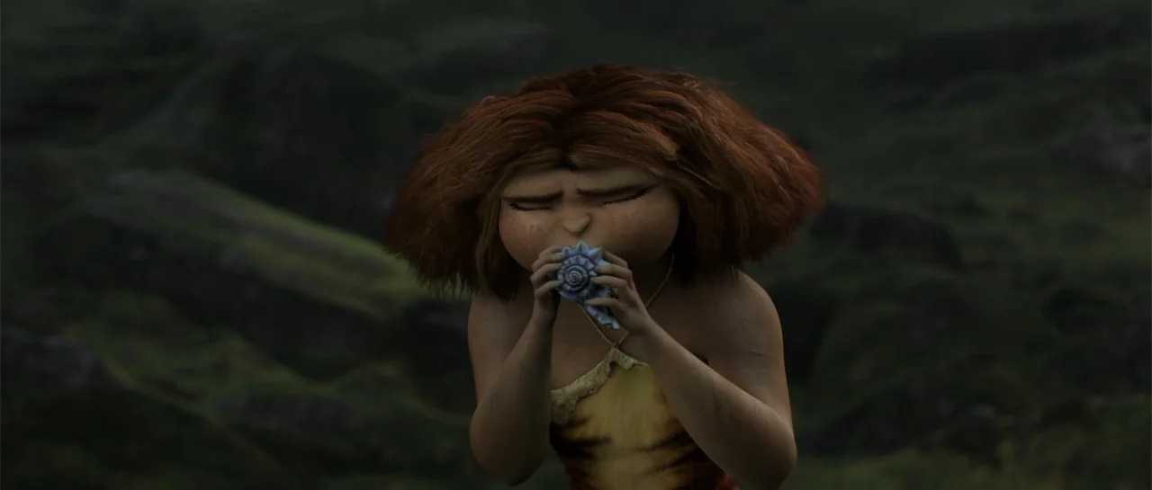 The Croods (2013) movie still: medium — A young girl with wild hair stands against a dark, out-of-focus rocky landscape. She holds a blue conch…
