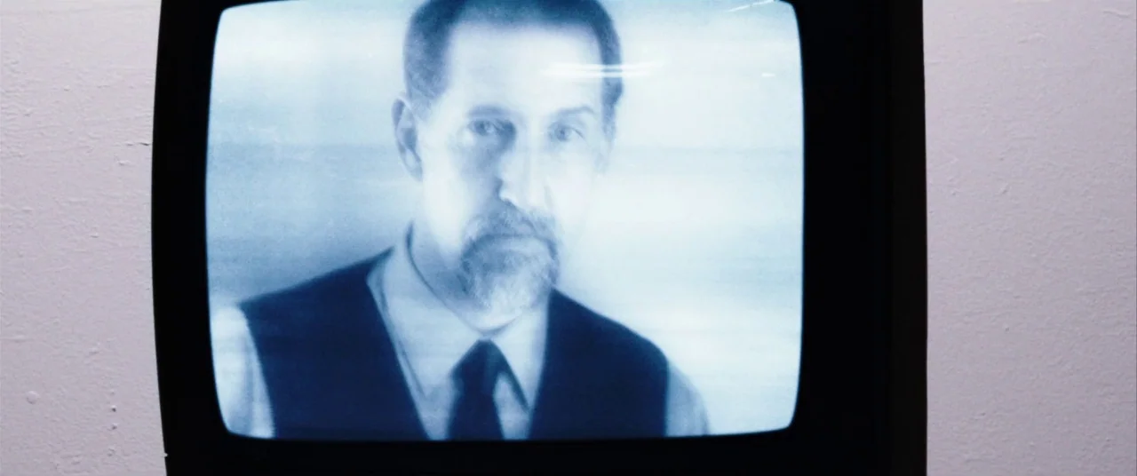 The Mind’s Eye (2015) movie still: closeup — A close-up of a middle-aged man displayed on a vintage television monitor. The image has a blue…