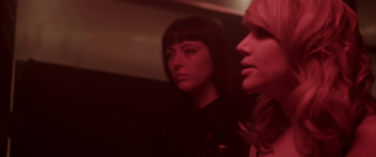 American Mary (2012) movie still: medium — Two women stand together in a dark space lit by deep red light. The women look toward the right of the…