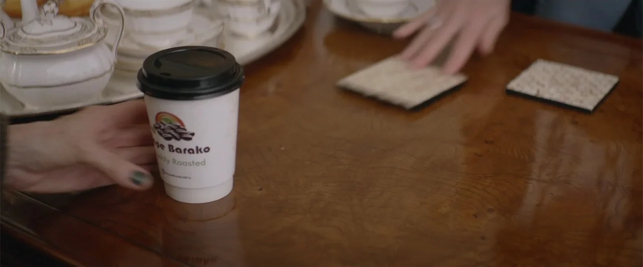 Hampstead (2017) movie still: medium — A polished wooden table holds a paper coffee cup and a porcelain tea service. Two pairs of hands move items…