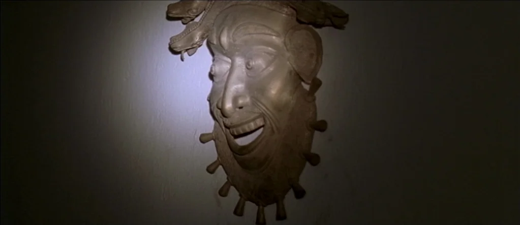 White Material (2009) movie still: closeup — A wall in a dimly lit room holds a decorative bronze mask. The mask features a grinning face with…