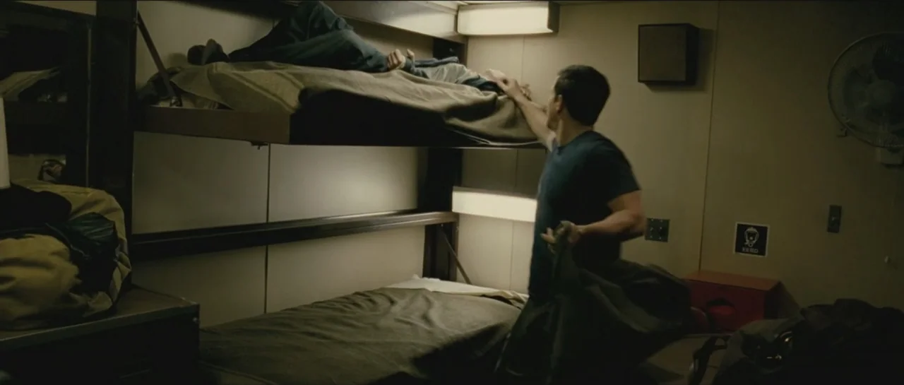 Contraband (2012) movie still: medium — A utilitarian bunk room with metal bunk beds and wall-mounted light fixtures. A man in a blue t-shirt…