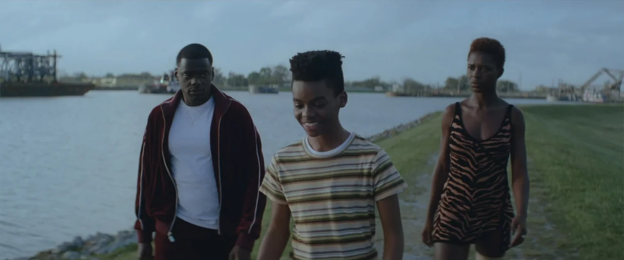Queen & Slim (2019) movie still: fullbody — A grassy embankment sits alongside a wide river with large industrial equipment visible far away. Two…