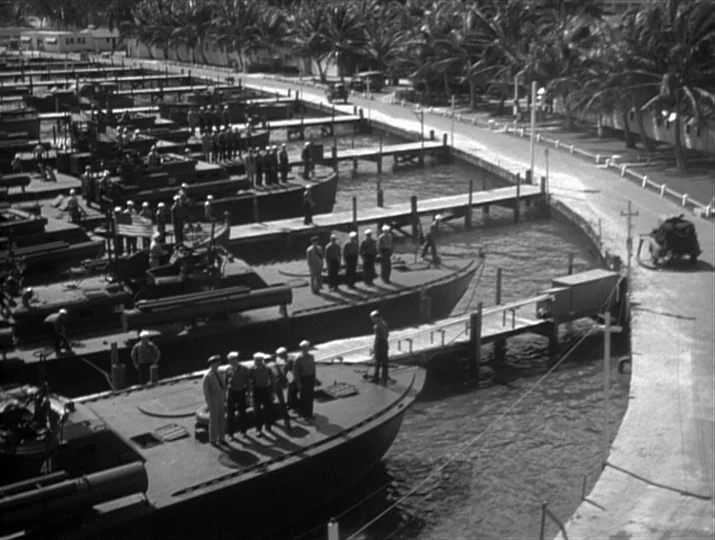 They Were Expendable (1945) movie still: wide — A military marina features several patrol boats lined up along wooden piers. Numerous sailors in…