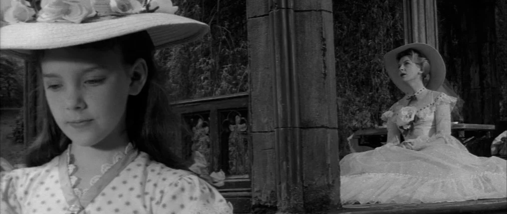 The Innocents (1961) movie still: medium — A wooden gazebo stands in a garden area with overgrown foliage in the background. A young girl is in the…