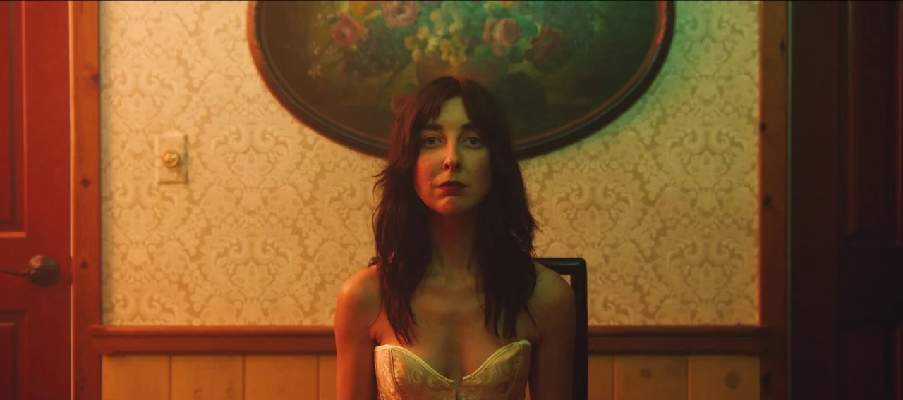 Fuzzy Head (2023) movie still: medium — A woman sits in front of a wall with patterned wallpaper and a floral painting. She is positioned centrally…
