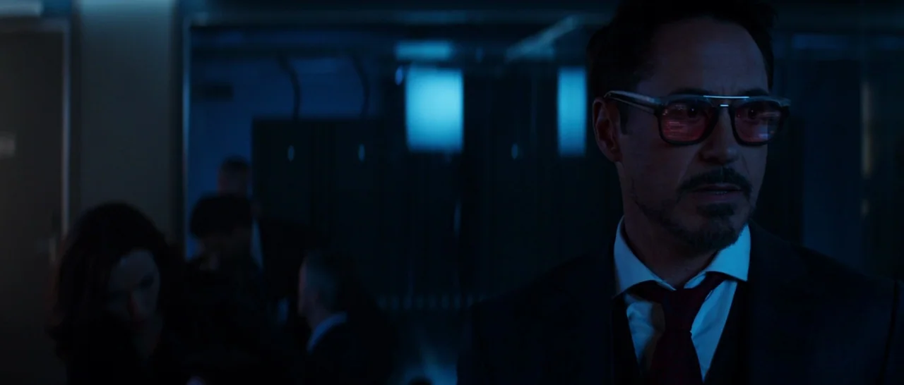 Captain America: Civil War (2016) movie still: medium — A dark, dimly lit corridor features blurred figures in the background near a glass wall. A…