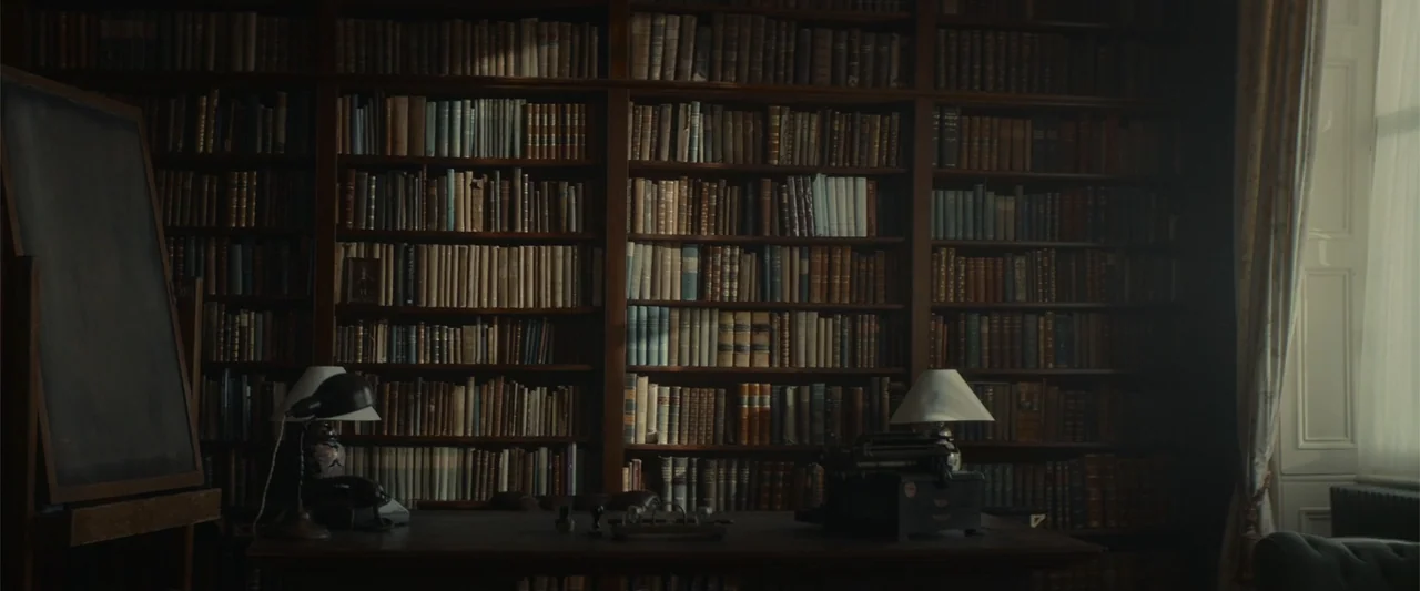The Cellar (2022) movie still: establishing — A wood-paneled room features floor-to-ceiling bookshelves packed with antique books and a central…