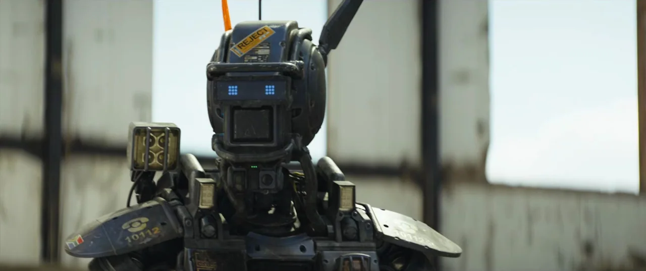 Chappie (2015) movie still: closeup — A robotic figure stands in front of a dilapidated structure with open window frames. The machine is heavily…