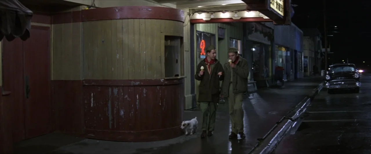 This Boy's Life (1993) movie still: fullbody — Two teenagers in scout uniforms walk down a quiet nighttime sidewalk past a theater. The boys are…