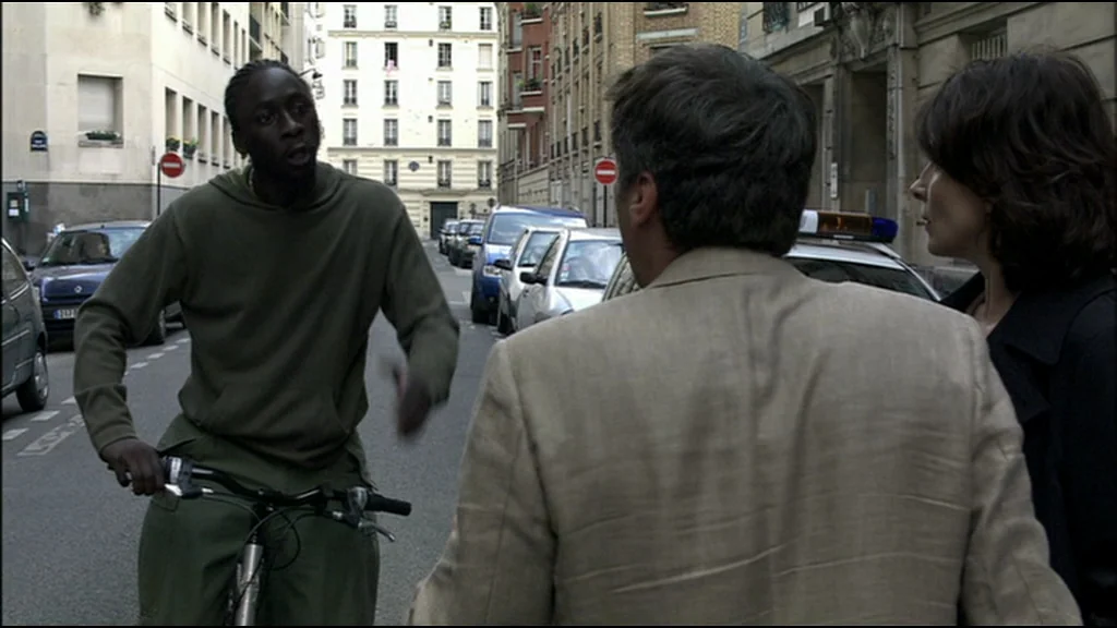 Caché (Hidden) (2005) movie still: medium — A Paris street with mid-rise apartment buildings and parked cars lining the curb. A man on a bicycle…
