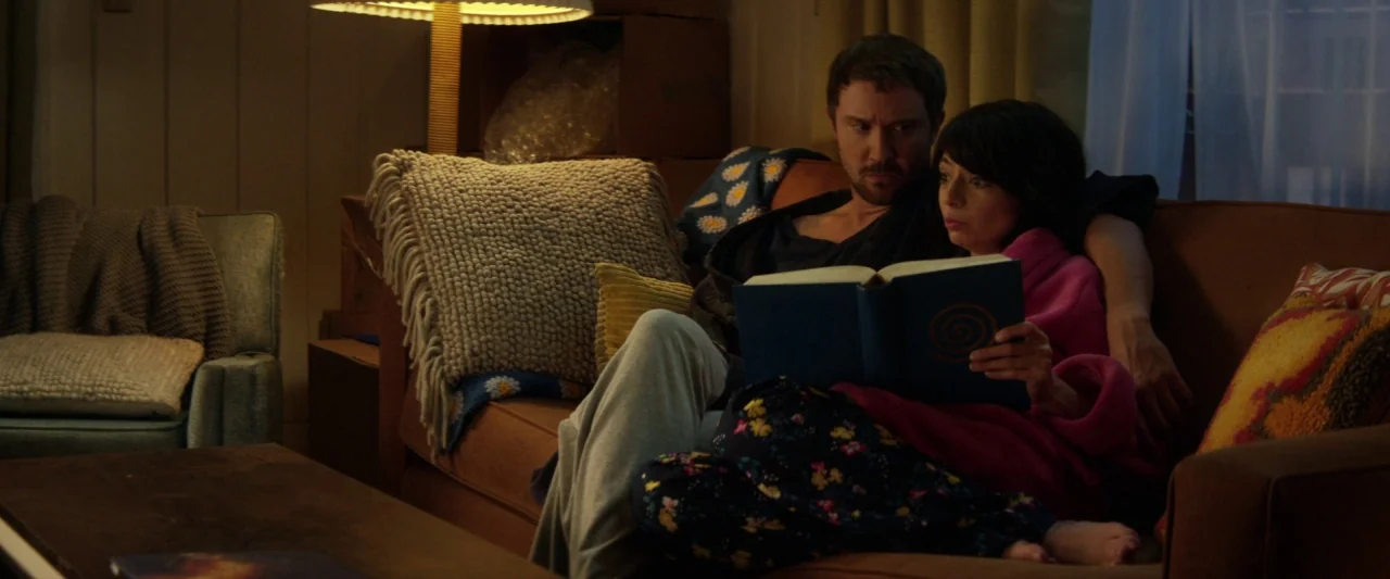 Seven Stages to Achieve Eternal Bliss (2018) movie still: medium — A cozy living room at night features a brown sofa with decorative pillows. A man…