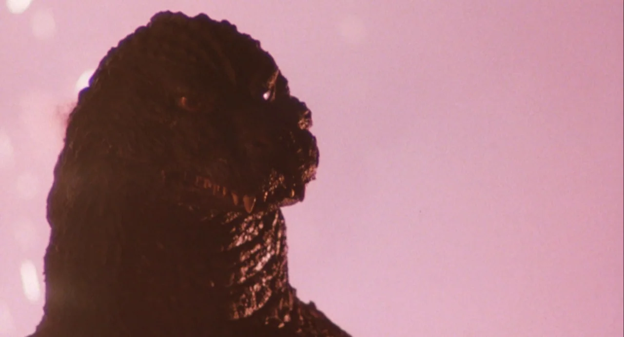 Godzilla and Mothra: The Battle for Earth (1992) movie still: closeup — A dark, scaly reptilian head is positioned against a vibrant, hazy pink…