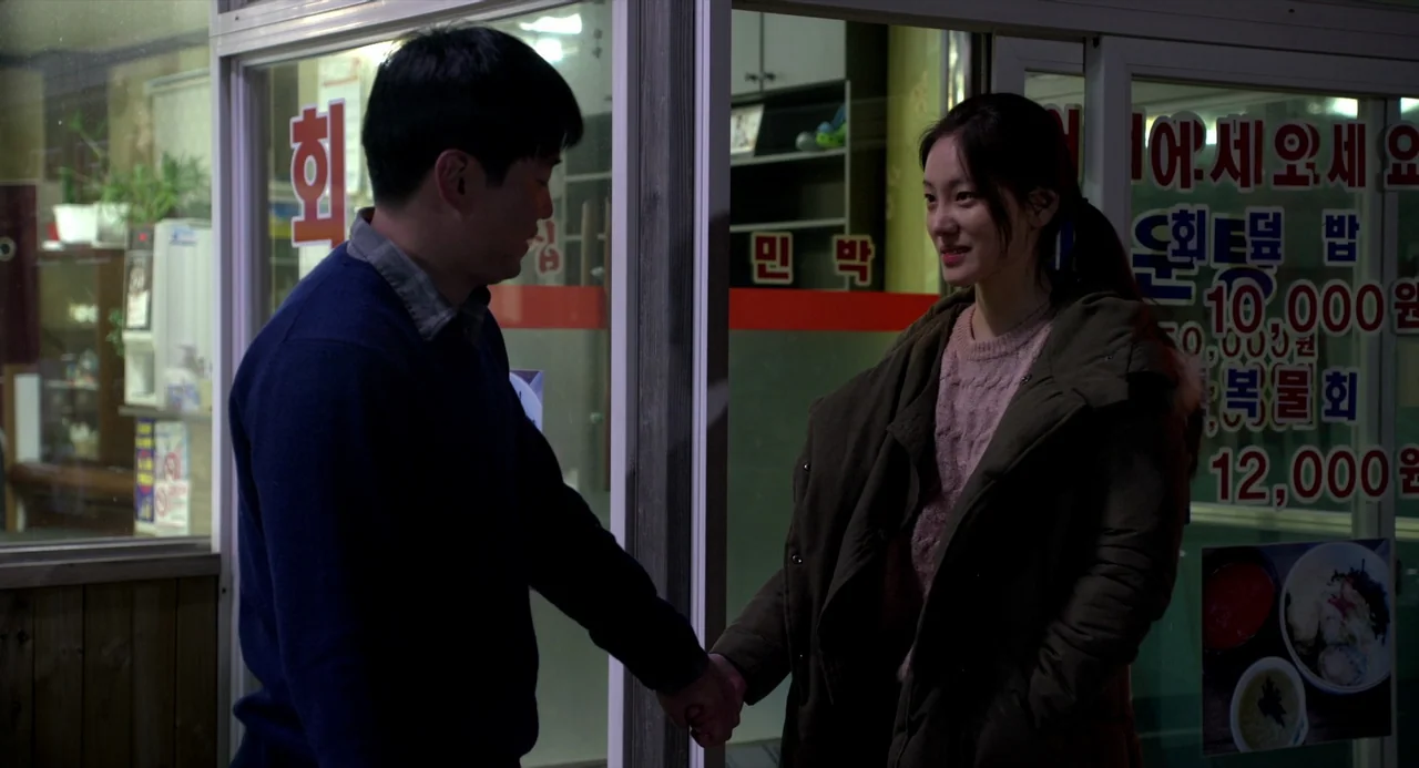 On The Beach At Night Alone (2017) movie still: medium — A man and a woman stand outside a glass-fronted storefront with Korean text signs. They…