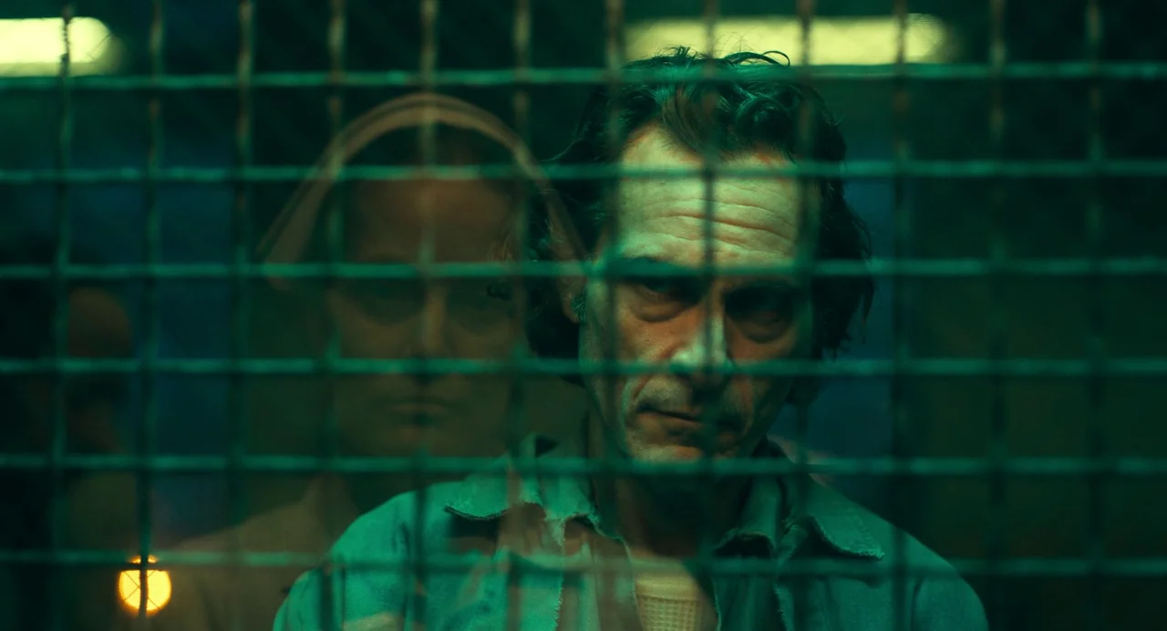 Joker: Folie à Deux (2024) movie still: closeup — A dimly lit industrial environment with a prominent metal mesh screen in the foreground. A man and…