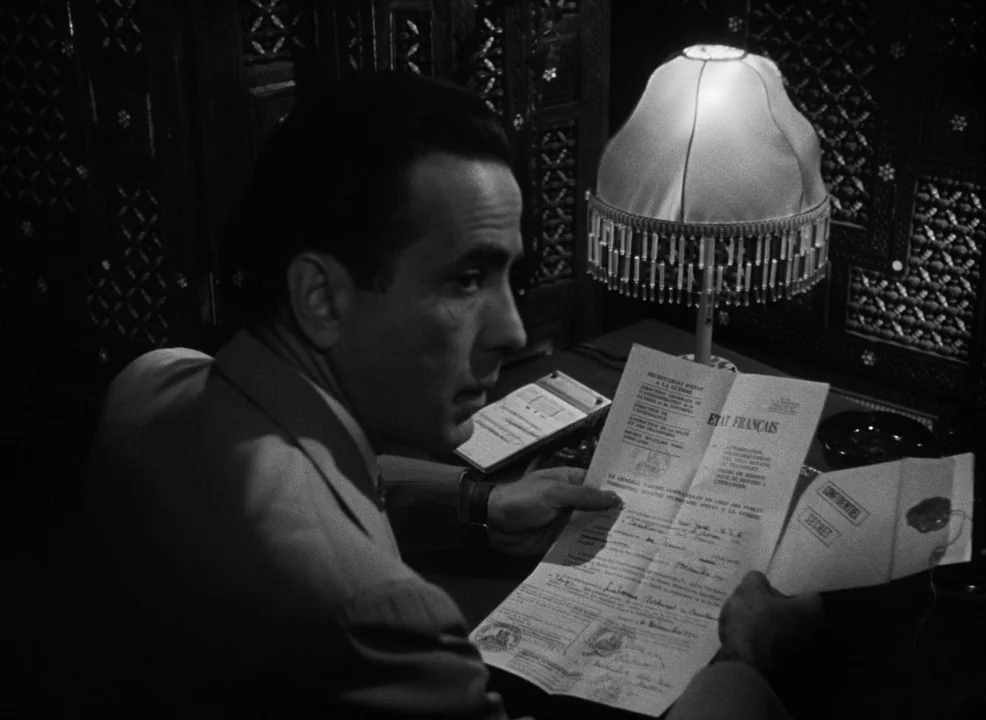 Casablanca (1942) movie still: medium — A dimly lit workspace with an ornate carved wooden partition in the background. A man sits at a table,…