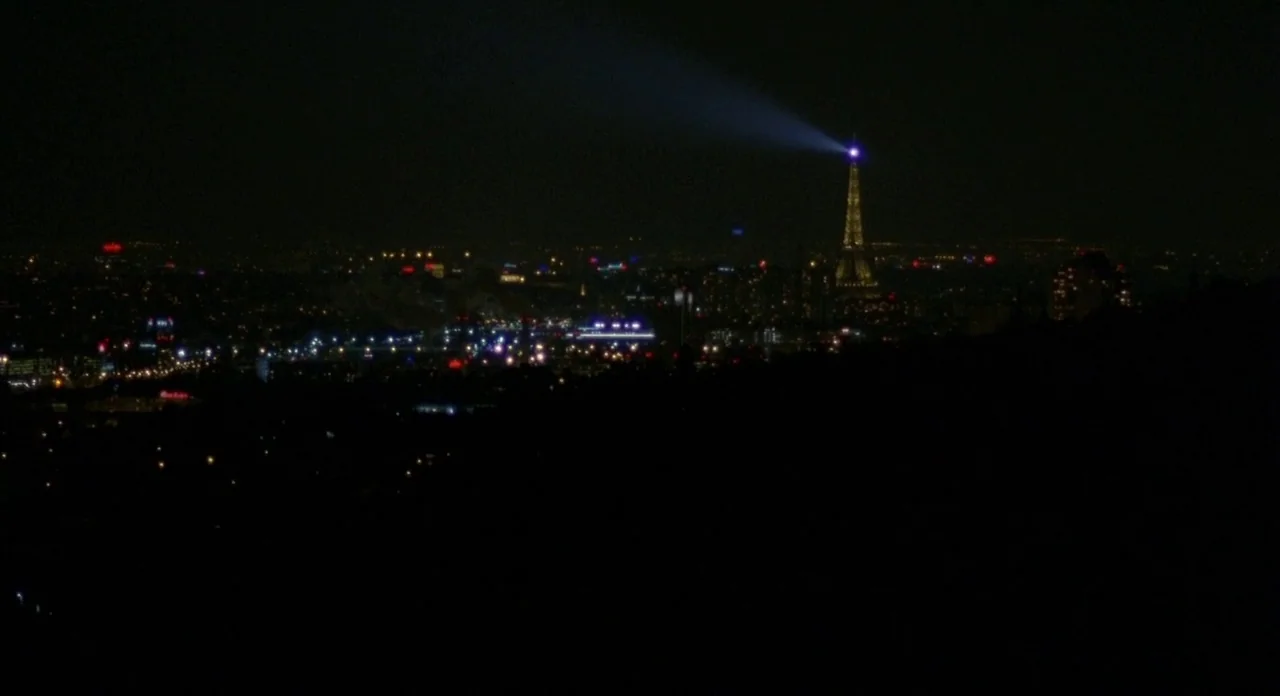 Read My Lips (2001) movie still: establishing — A panoramic night view of a distant city skyline under a dark sky. The Eiffel Tower stands…