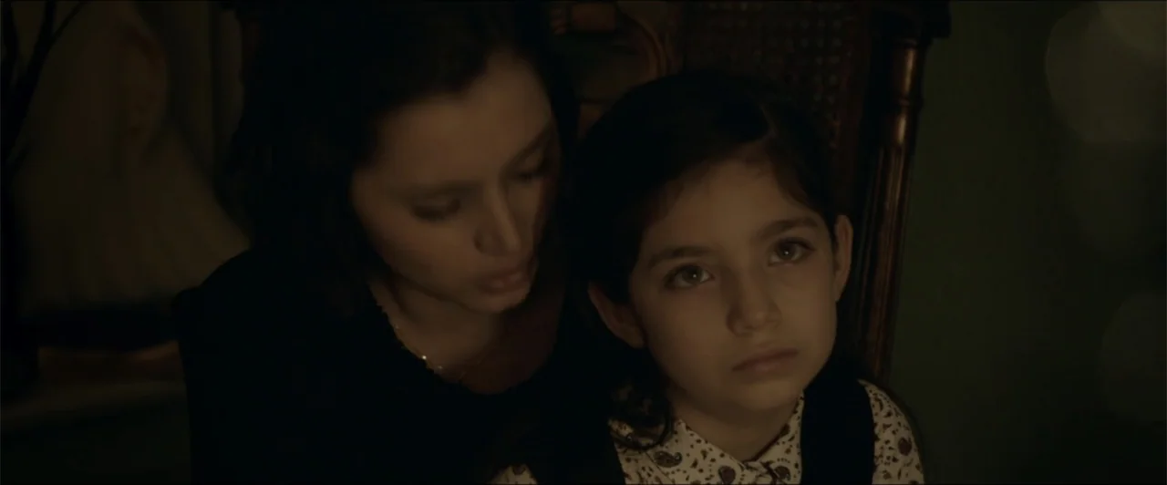 Exposed (2016) movie still: closeup — A dark indoor space features a young girl and a woman sitting closely together. The woman leans toward the…